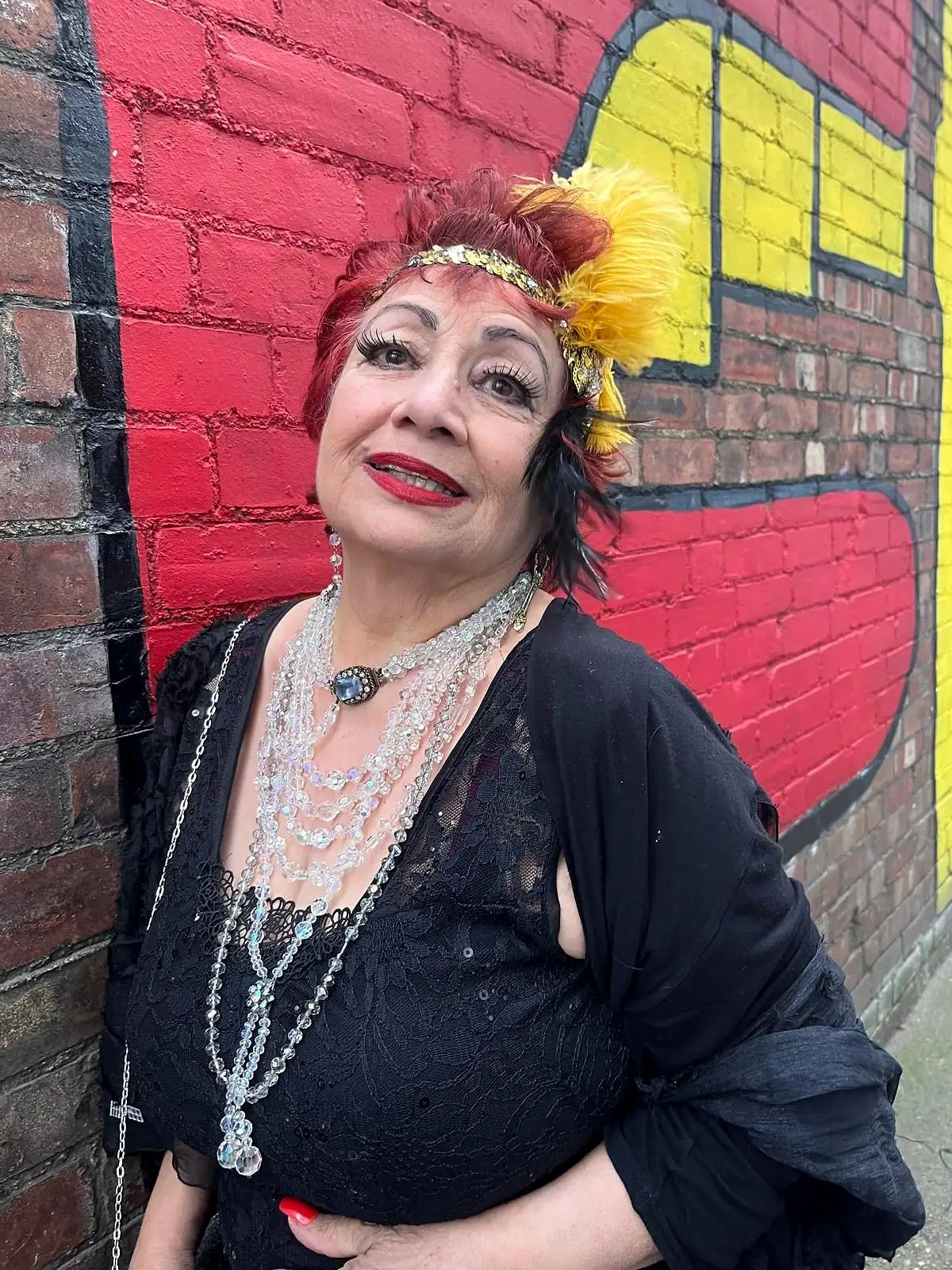 Anahita Eastwood in black lace with crystal necklaces, a sequinned headband and a yellow feather, in front of a red painted brick mural