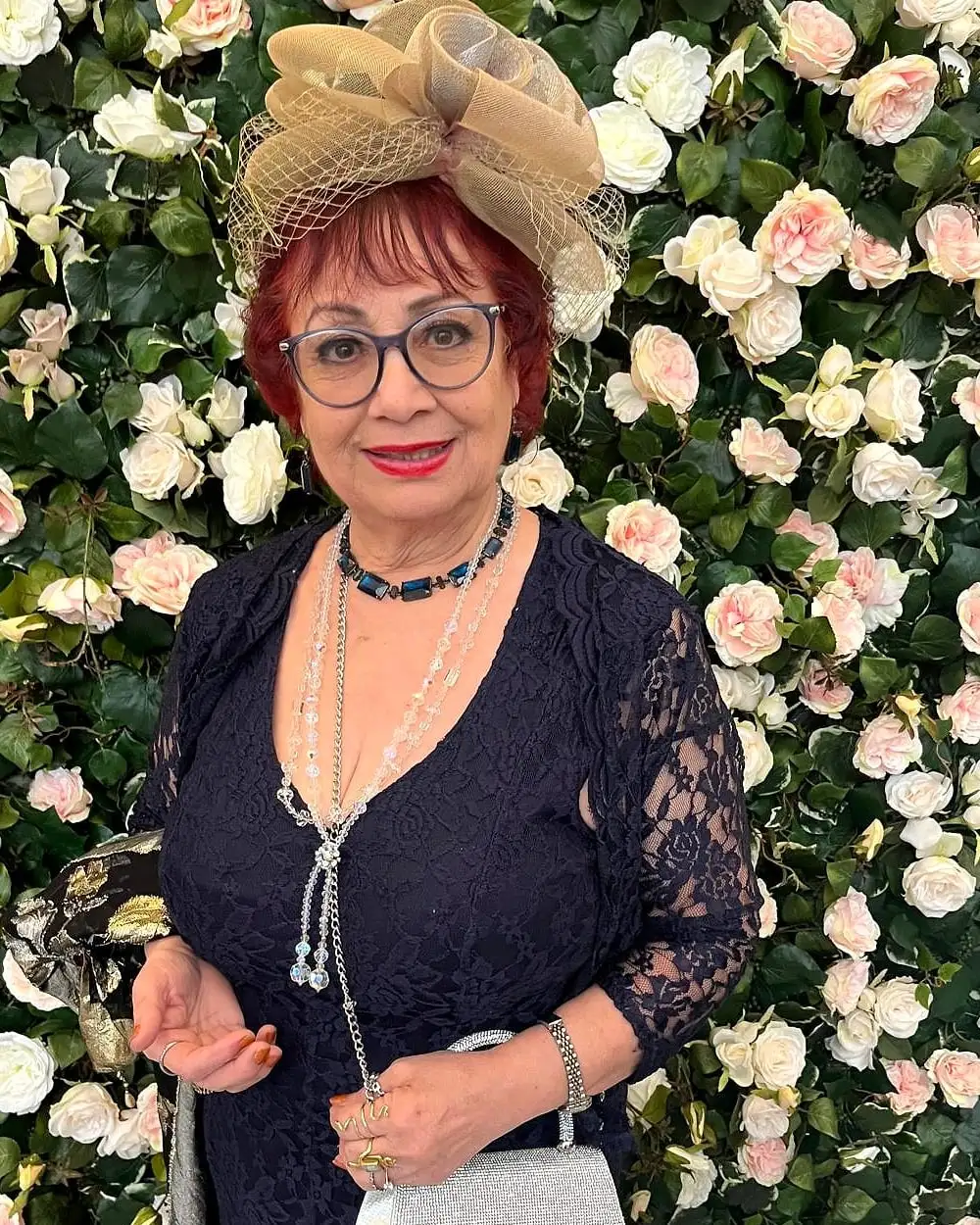 Anahita Eastwood in a navy lace dress and a fascinator, smiling in front of a wall of pale pink and white roses