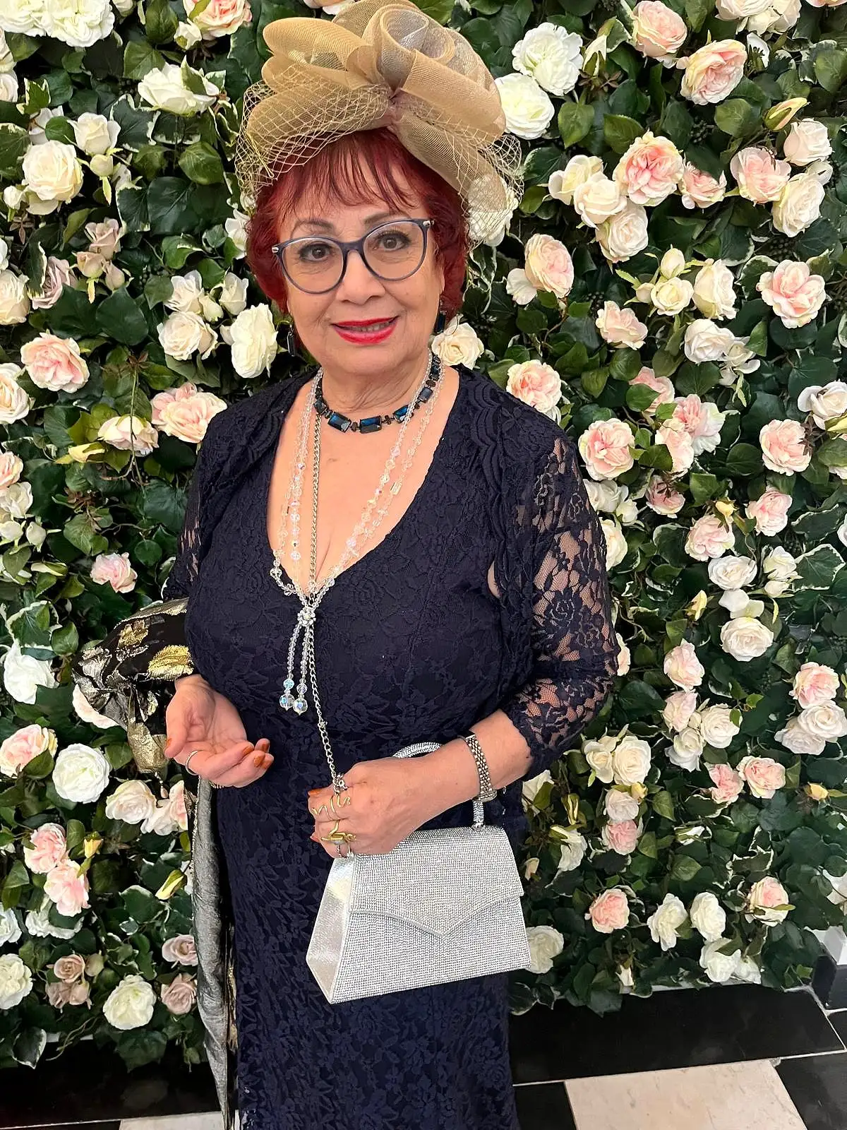 Anahita Eastwood in a navy lace dress and a fascinator, smiling in front of a wall of pale pink and white roses