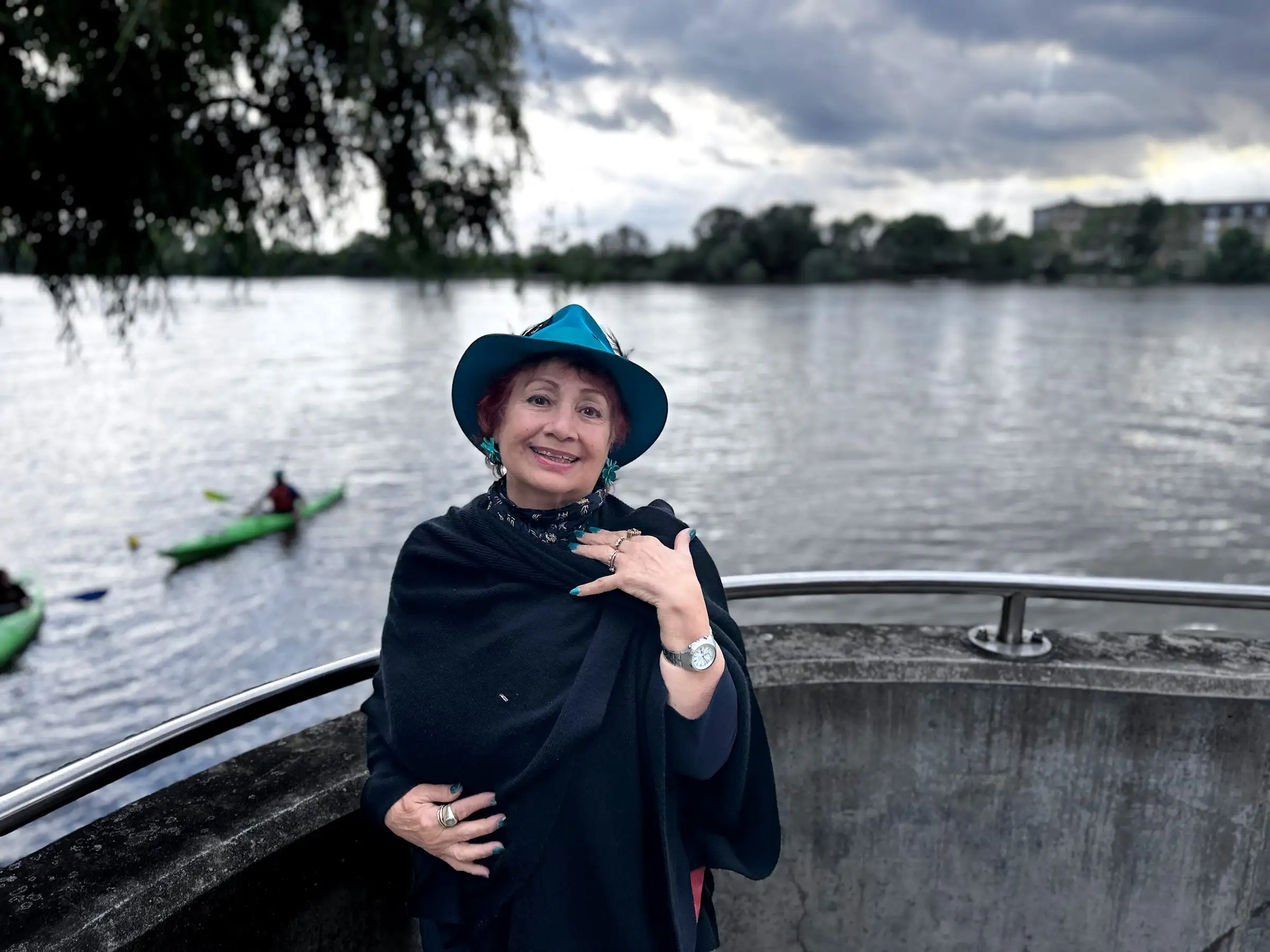 Anahita Eastwood in a teal hat and a dark wrap, smiling by a wide river under a grey sky, kayaks passing behind her