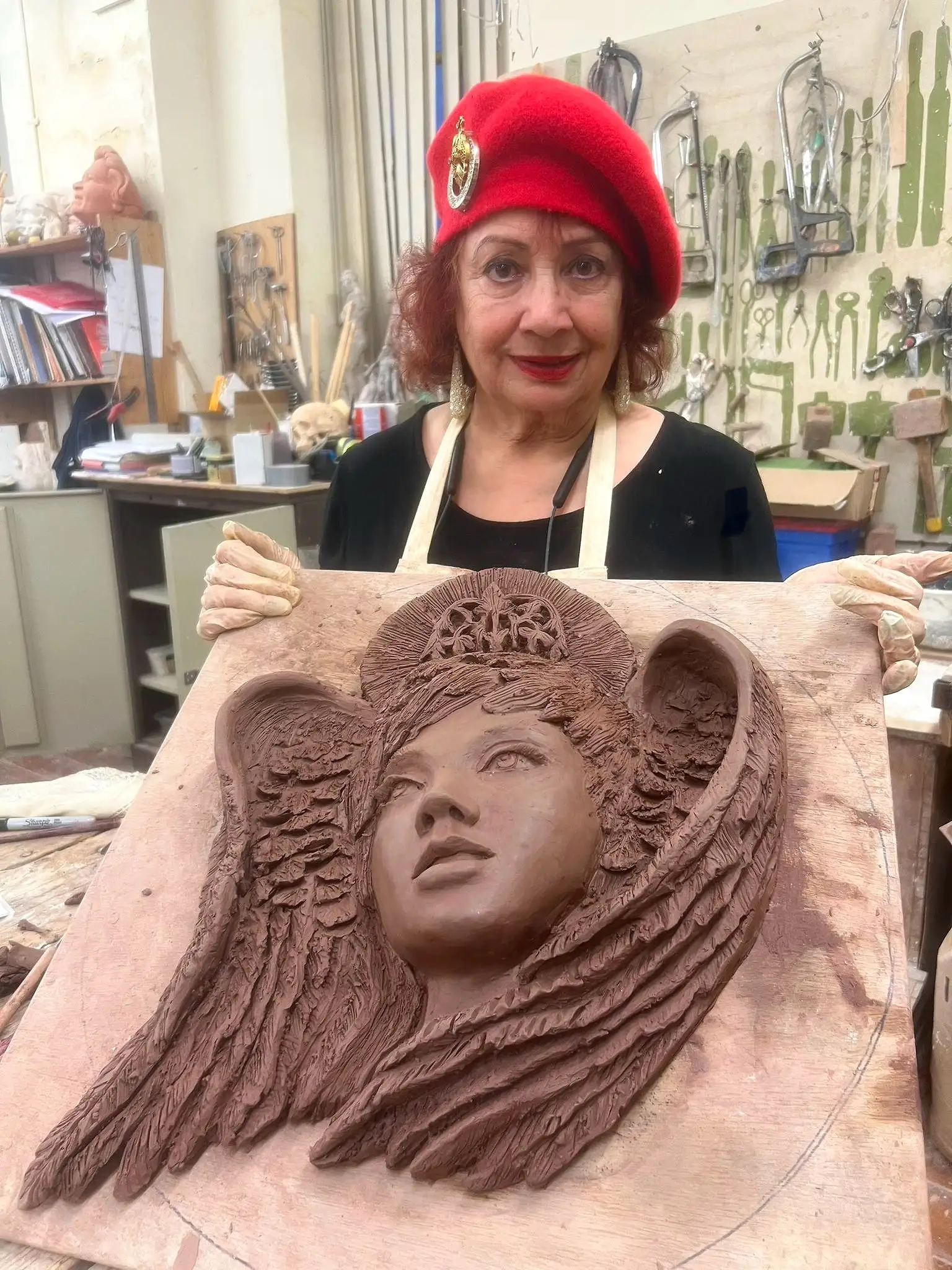 Anahita Eastwood in a red beret and an apron in a sculpture studio, holding up a clay relief of a winged face in progress