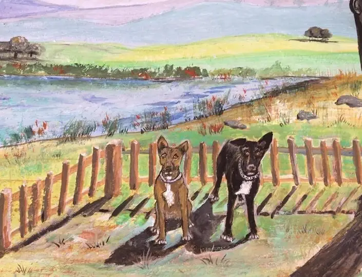 Two dogs, one tawny brown and one black, sit side by side at a wooden paling fence with a wide river, green meadows and pale blue hills stretching behind.