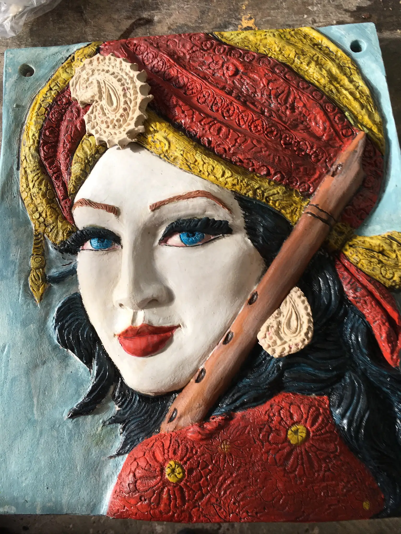 A glazed ceramic wall relief of a young woman with pale skin, blue eyes and red lips, wearing a red and yellow paisley turban and a red flower patterned garment, holding a wooden ney flute across her chest, mounted on a turquoise slab