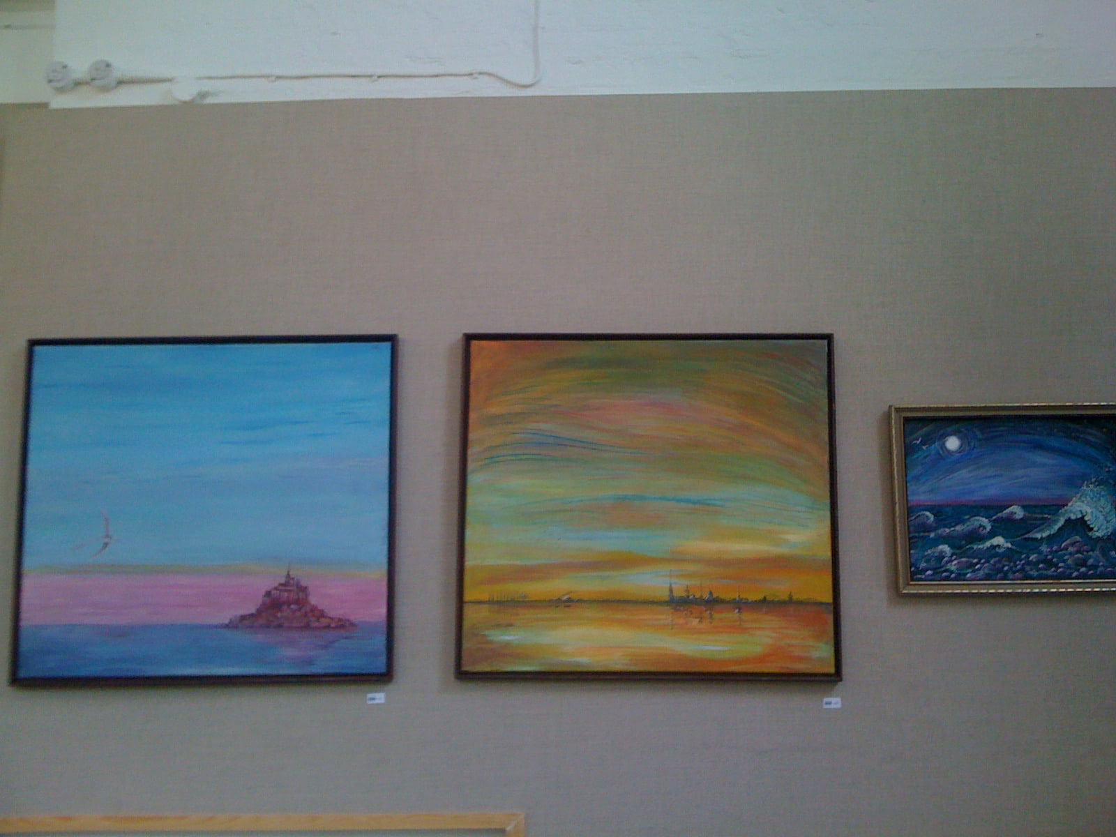 Three of Anahita's seascapes hung together on a taupe gallery wall: an island fortress under a blue sky at dawn, a warm sunset over calm water and a small moonlit night sea