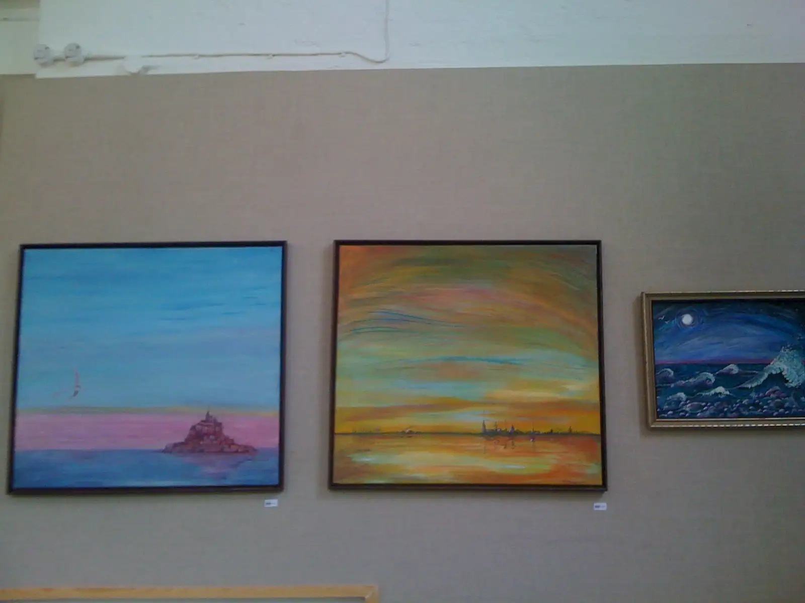 Three of Anahita's seascapes hung together on a taupe gallery wall: an island fortress under a blue sky at dawn, a warm sunset over calm water and a small moonlit night sea