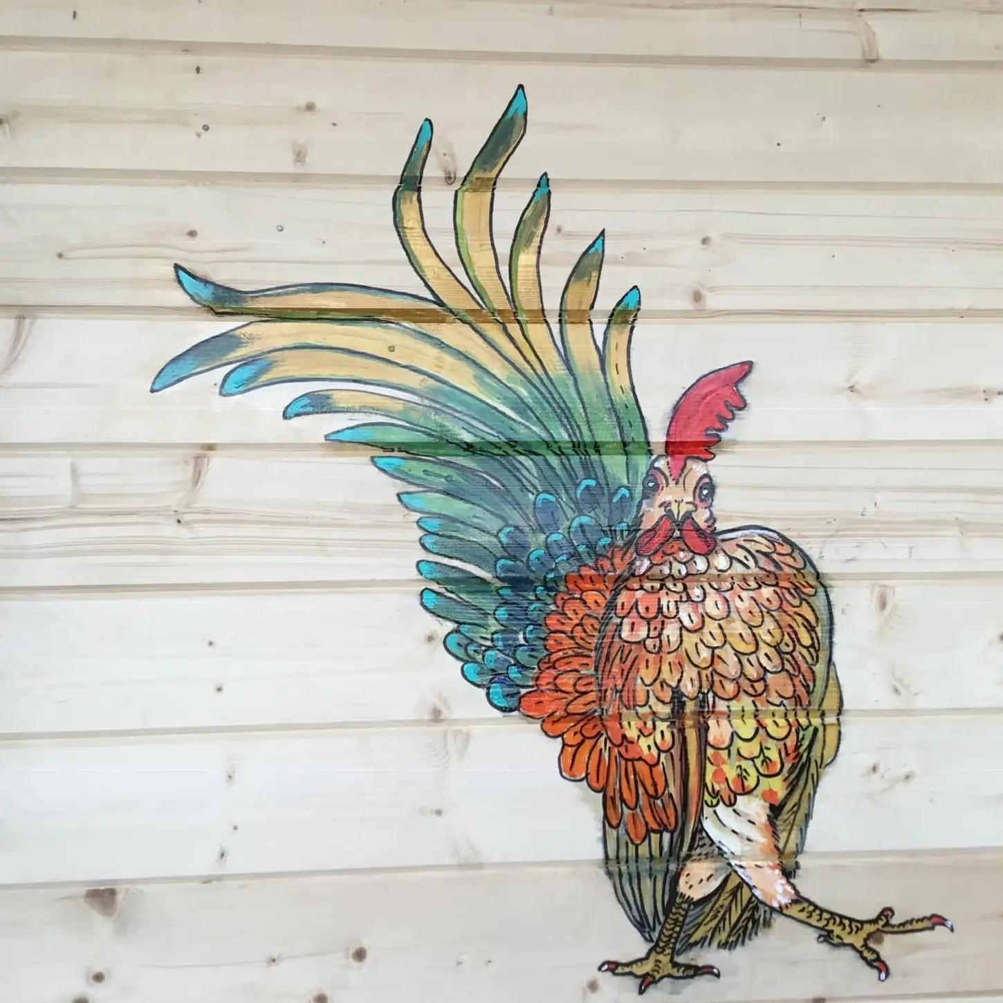 A proud cockerel with a red comb, orange and gold body feathers and long sweeping teal, green and yellow tail feathers, painted directly onto pale pine cladding.