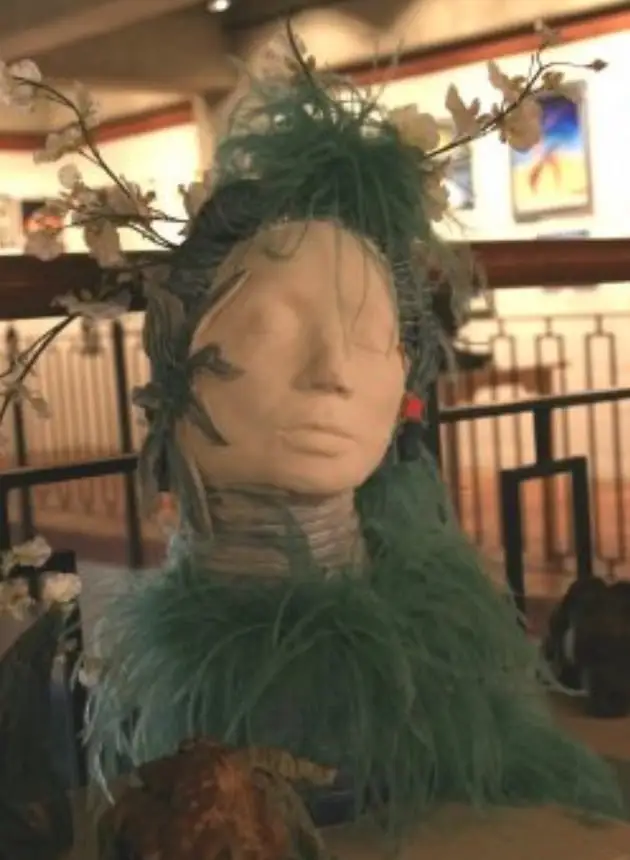 A pale ceramic head of a woman with closed eyes crowned by dark teal leaves and white blossom branches, a soft teal green collar around the shoulders, on a gallery plinth