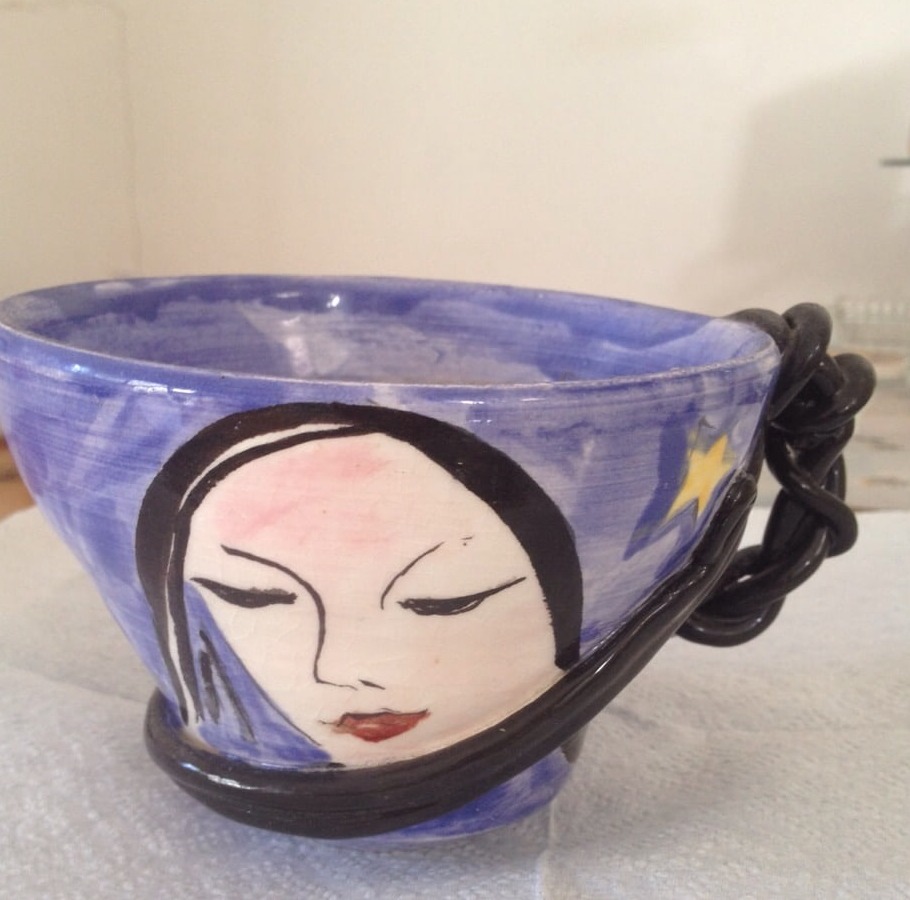 A cornflower blue glazed ceramic cup with a painted face of a dark-haired woman with closed eyes and red lips, a yellow shooting star on the reverse and a thick coiled black handle
