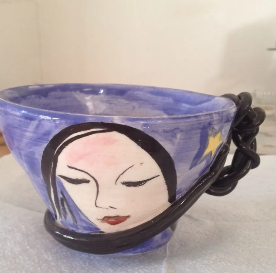 A cornflower blue glazed ceramic cup with a painted face of a dark-haired woman with closed eyes and red lips, a yellow shooting star on the reverse and a thick coiled black handle