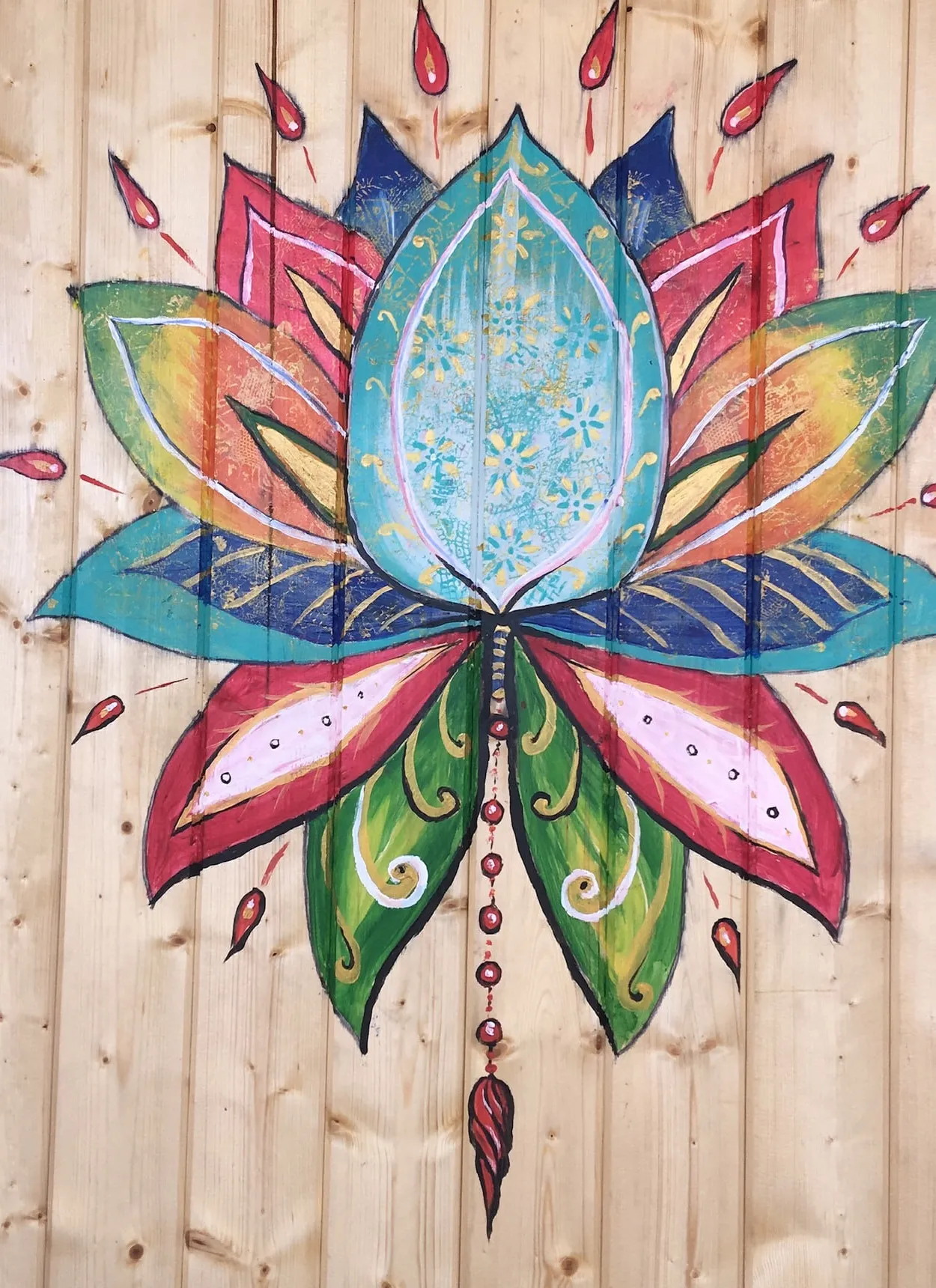 Bright stylised lotus painted on pale pine cladding, its petals layered in turquoise, cobalt, coral, rose, green and gold with a hanging string of red drop-shaped beads.