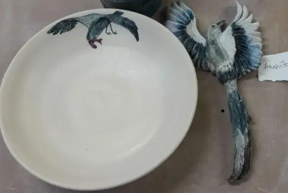 A cream ceramic bowl with a hand-painted blue-grey pigeon in flight sits beside a companion ceramic bird with fully spread wings and a long trailing tail on a workbench