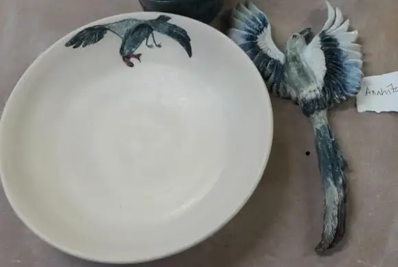 A cream ceramic bowl with a hand-painted blue-grey pigeon in flight sits beside a companion ceramic bird with fully spread wings and a long trailing tail on a workbench