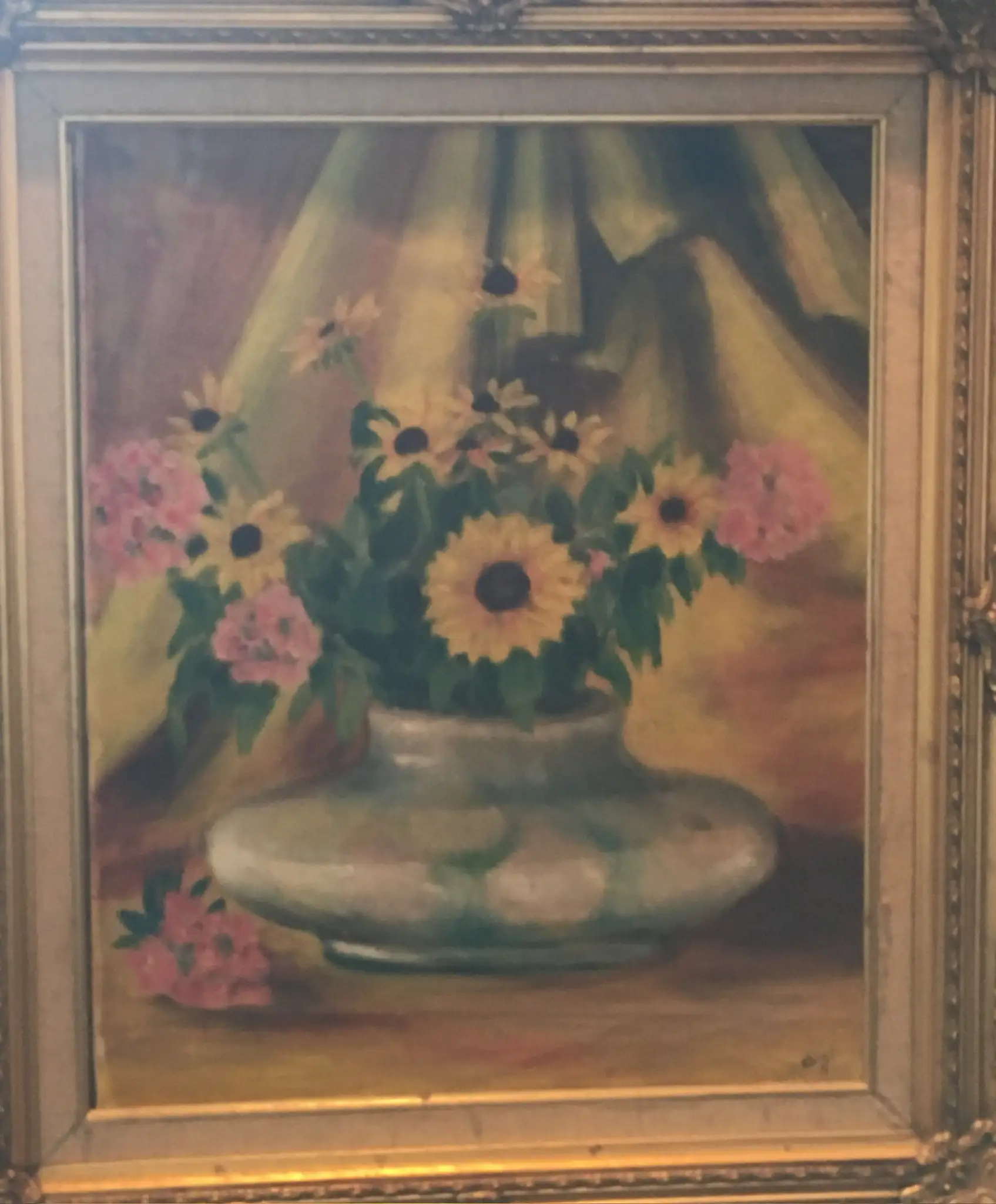A still life of yellow daisies with black centres and small pink flowers in a squat green-grey glazed vase, set against draped green and rose fabric, in an ornate gilt frame