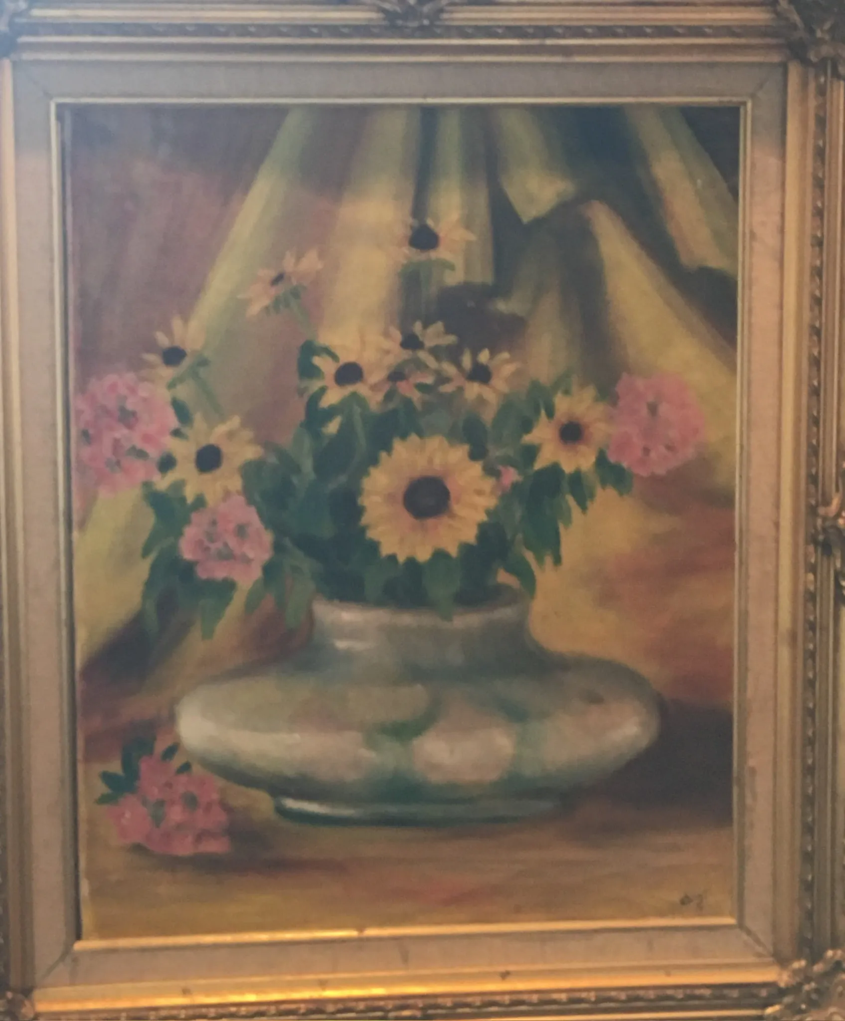 A still life of yellow daisies with black centres and small pink flowers in a squat green-grey glazed vase, set against draped green and rose fabric, in an ornate gilt frame