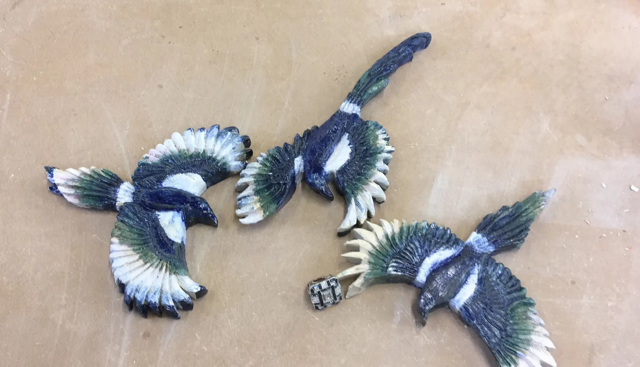 Three glazed ceramic magpie wall reliefs with outstretched wings in cobalt blue, iridescent green and white, arranged on a studio board.