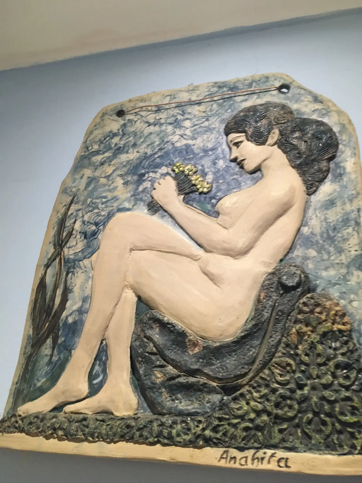 A glazed ceramic wall relief of a seated female nude in profile holding a small posy of yellow flowers, her pale cream body raised against a mottled cobalt and green background with a foamy green base.