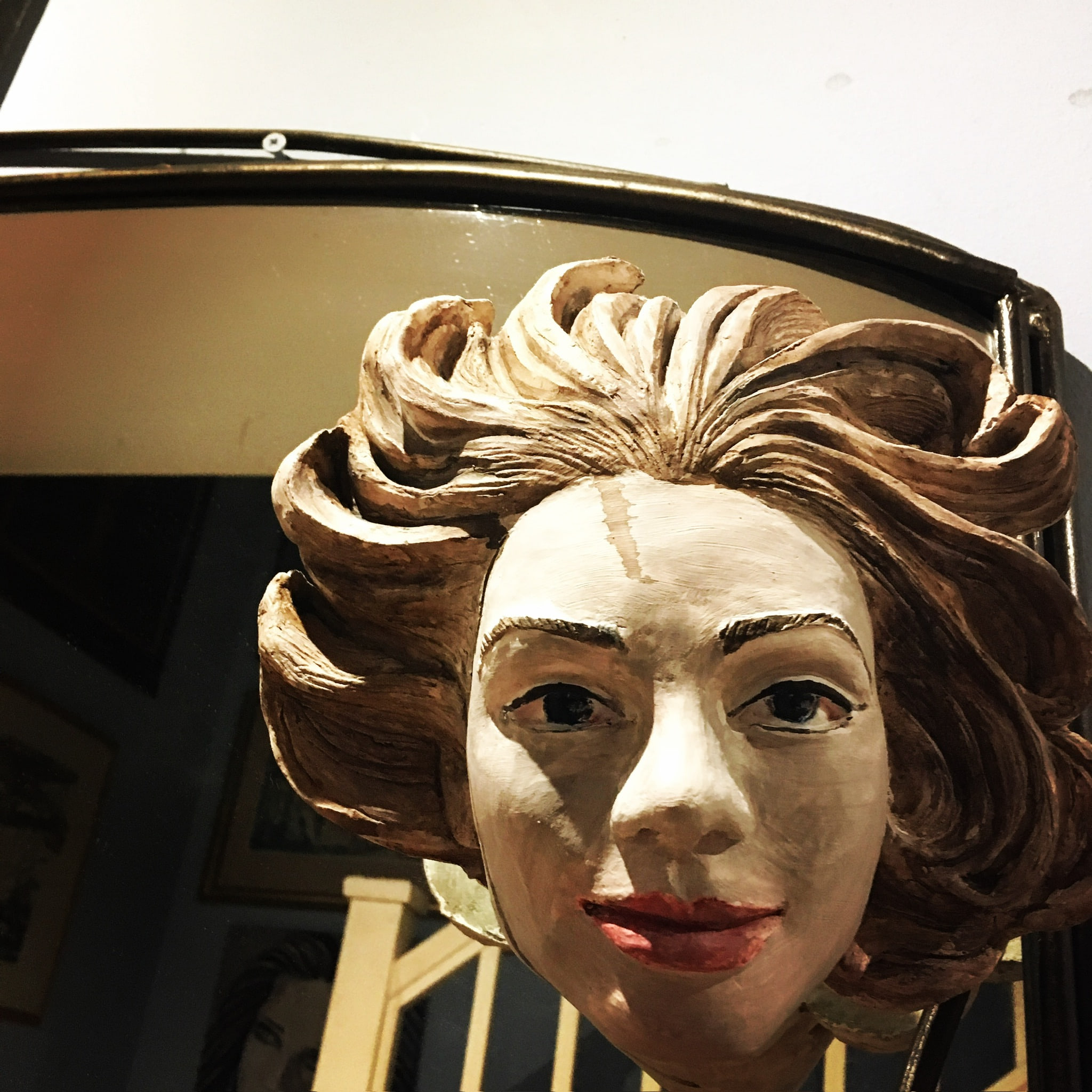 A glazed ceramic head of a smiling woman with sweeping auburn hair, arched brows, bright blue eyes and red lips, mounted above a gilt oval mirror.