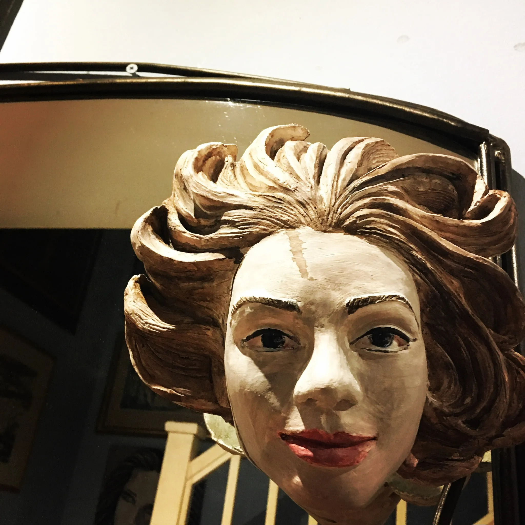 A glazed ceramic head of a smiling woman with sweeping auburn hair, arched brows, bright blue eyes and red lips, mounted above a gilt oval mirror.